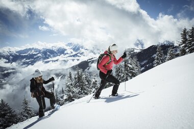 Two people snowshoeing in a snowy mountain landscape with clouds and mountains in the background. | © Zell am See-Kaprun Tourismus