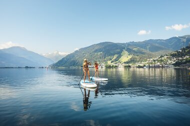 Two people stand-up paddleboarding on Lake Zeller with mountains in the background on a sunny day. | © Zell am See-Kaprun Tourismus