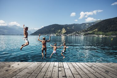 Children jump and splash in Lake Zeller on a sunny day, surrounded by mountains and a small town in the background. | © Korbinian Seifert