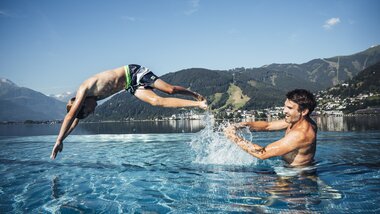 Two people in the lake, one jumping into the water while the other stands in the water greeting, surrounded by mountains and sky. | © Zell am See-Kaprun Tourismus