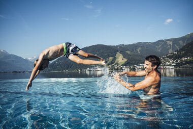 Two people in the lake, one jumping into the water while the other stands in the water greeting, surrounded by mountains and sky. | © Zell am See-Kaprun Tourismus