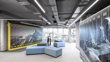 Modern ski storage with yellow locker units and mountain images on the walls, comfortable seating in the center, large windows with a view outside. | © Wöckinger
