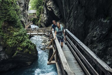 Hiker walking on a narrow wooden bridge beside a river in Sigmund-Thun Gorge near Kaprun, surrounded by tall rock walls and lush trees. | © Zell am See-Kaprun Tourismus