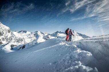 Two skiers on a snowy mountain ridge with peaks in the background and a clear sky. | © Kitzsteinhorn
