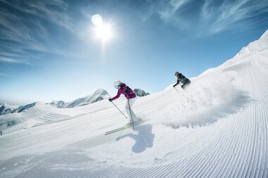Two skiers on a groomed slope at Kitzsteinhorn under sunshine and a clear blue sky. | © Kitzsteinhorn