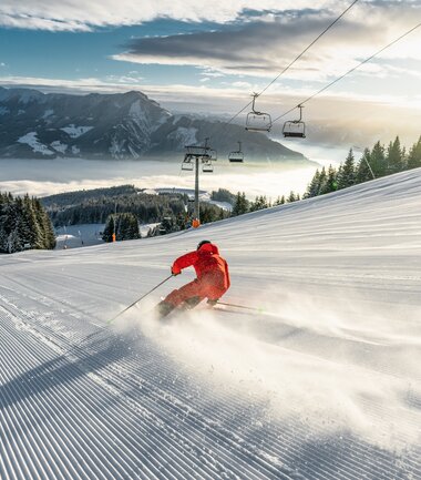 Skier skier descending a snowy slope at sunrise with a mountain landscape, ski lift, and pine trees in the background. | © Zell am See-Kaprun Tourismus