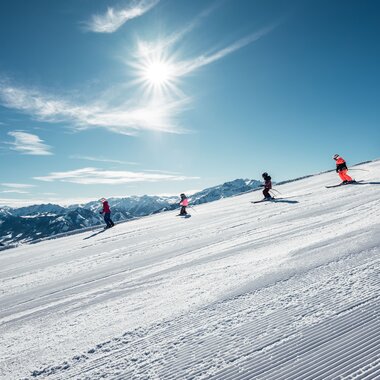 Skiers in the sun on a snowy slope with mountains in the background. | © Zell am See-Kaprun Tourismus