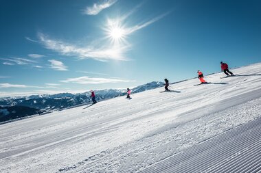 Skiers in the sun on a snowy slope with mountains in the background. | © Zell am See-Kaprun Tourismus