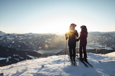 Two skiers on Schmittenhöhe at sunrise, surrounded by snow-covered mountains. | © Zell am See-Kaprun Tourismus