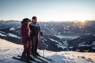 Two skiers on a snow-covered slope with mountain and lake scenery in the sunrise background. | © Zell am See-Kaprun Tourismus