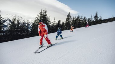 Ski instructor with children skiing on a snowy slope, surrounded by pine trees. | © Zell am See-Kaprun Tourismus