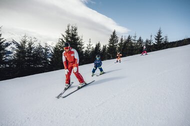 Ski instructor with children skiing on a snowy slope, surrounded by pine trees. | © Zell am See-Kaprun Tourismus