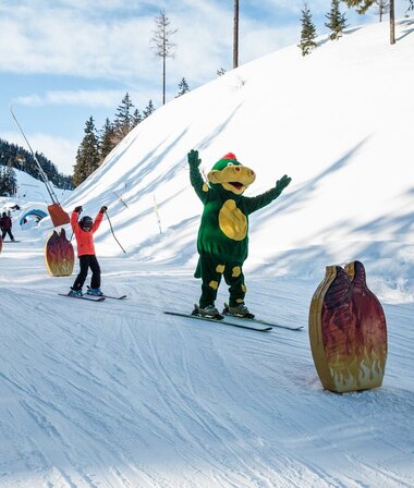 Skiers on a snowy slope with a dinosaur-costumed character performing at a dragon park. | © Schmittenhöhe