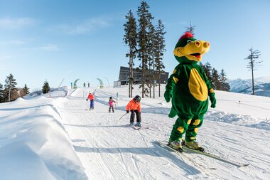 Children on skis on a snowy slope with a Skimountain mascot in a costume and ski instructor. | © Zell am See-Kaprun Tourismus