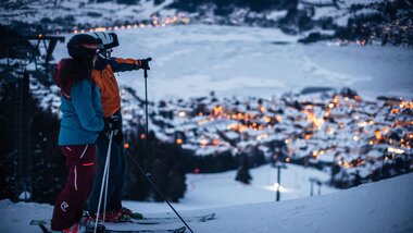 Skier with helmets and gear at night, overlooking a lit-up town in a snowy valley. | © Christian Mairitsch