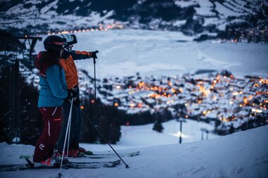 Skier with helmets and gear at night, overlooking a lit-up town in a snowy valley. | © Christian Mairitsch