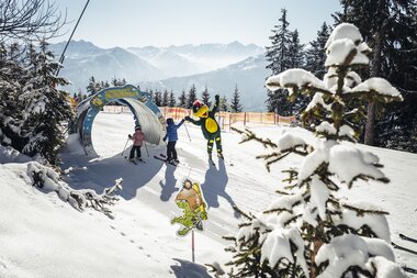 Children skiing and playing on the snowy slope with a guide and another skier in the background, surrounded by snow-covered trees. | © Zell am See-Kaprun Tourismus