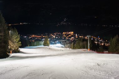 Nighttime view of a illuminated ski slope on Schmittenhöhe with a town in the distance. | © Christian Mairitsch