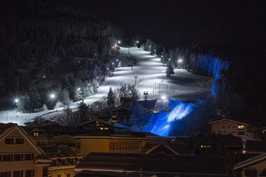 Lights on a nighttime ski slope in a snowy mountain area, surrounded by trees and buildings. | © Schmittenhöhe
