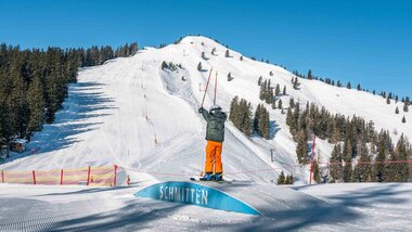 Skier at the beginning of the slope with a fun park and snow-covered hill on the Schmittenhöhe, surrounded by trees under a blue sky. | © Schmittenhöhe