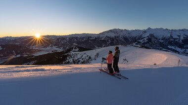 Skier on a snowy mountain during sunset with a mountain range in the background. | © Schmittenhöhe