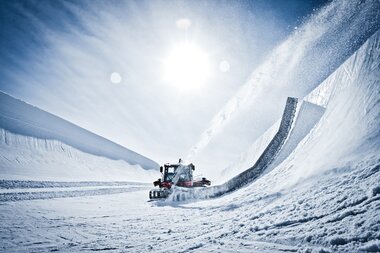 View of a snow-covered snowpark on a sunny day, with a snow grooming machine preparing the terrain. | © Kitzsteinhorn