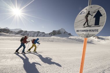 Skiers on a mountain trail under bright sunshine, with a sign indicating a ski touring route in Zell am See, Austria. | © Robert Niedring