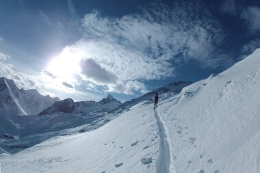 Ski tour on snowy slope with view of mountain peaks and cloudy sky, sunlight illuminating the landscape. | © Stephan Obenaus