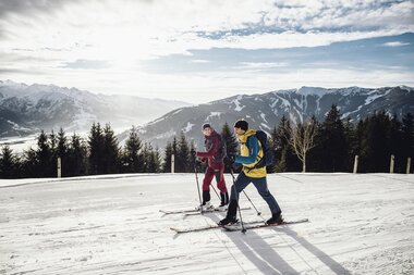 Two people ski touring in a snowy mountain landscape with mountains in the background and cloudy sky. | © Seifert