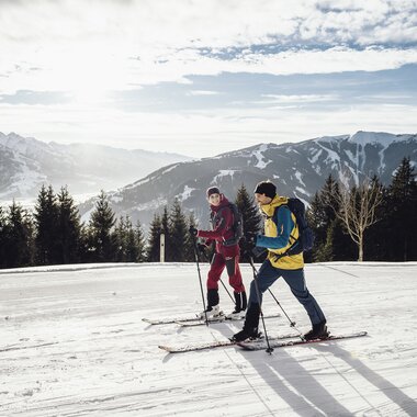 Two people ski touring in a snowy mountain landscape with mountains in the background and cloudy sky. | © Seifert