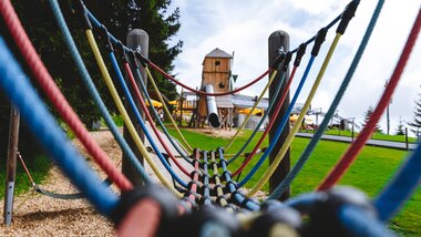 View through a playground net of a playground with trees and wooden huts in the background on a summer field. | © Schmittenhöhe, Alpine Vision