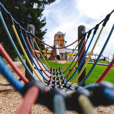 View through a playground net of a playground with trees and wooden huts in the background on a summer field. | © Schmittenhöhe, Alpine Vision