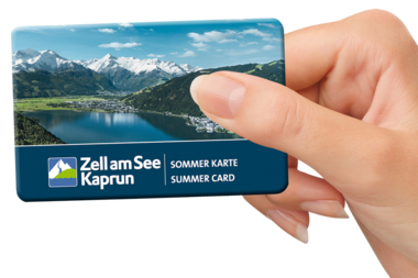 A hand holding a summer card for Zell am See-Kaprun with mountain and lake scenery in the background. | © Zell am See-Kaprun Tourismus