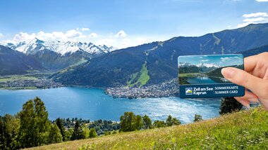 View of Lake Zell with the Alps in the background, someone holding a Summer Card for Zell am See-Kaprun. | © Zell am See-Kaprun Tourismus