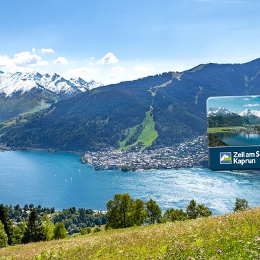 View of Lake Zell with the Alps in the background, someone holding a Summer Card for Zell am See-Kaprun. | © Zell am See-Kaprun Tourismus