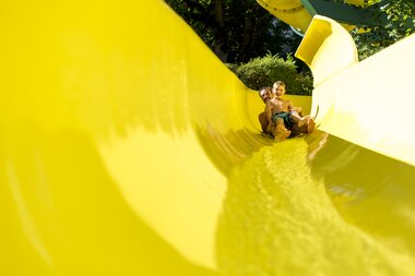 Child sliding down a large yellow water slide at the beach in Zell am See-Kaprun on a sunny day. | © Zell am See-Kaprun Tourismus