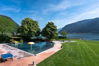 A swimming pool with sunbeds and an umbrella by a lake, surrounded by trees and mountains on a sunny day. | © Johannes Radlwimmer