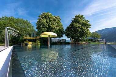 Outdoor swimming pool with water slide, trees, and mountain landscape in the background on a clear day. | © Johannes Radlwimmer
