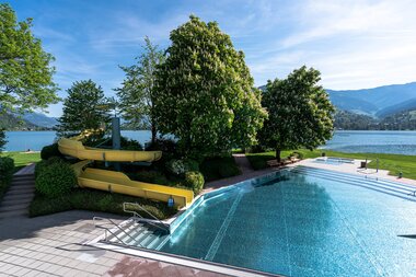 A swimming pool with water slides, trees, and a mountain view in the background, overlooking a lake. | © Johannes Radlwimmer