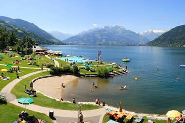 View of a lakeside beach area with loungers, umbrellas, and walking paths against a mountainous backdrop. | © FREGES Zell am See