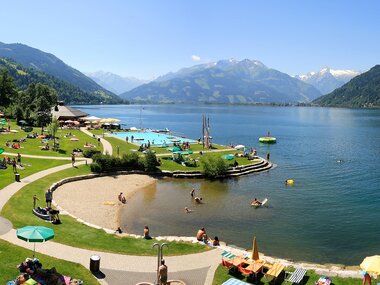 View of a lakeside beach area with loungers, umbrellas, and walking paths against a mountainous backdrop. | © FREGES Zell am See