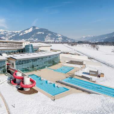 Aerial view of a winter hotel with outdoor pools and slides, surrounded by snow-covered landscape and mountains. | © Tauern SPA Zell am See-Kaprun