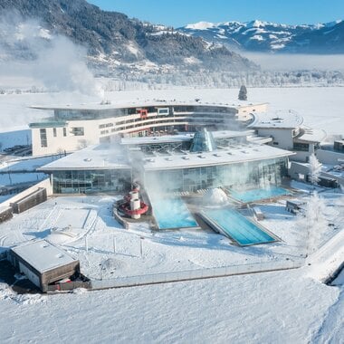Aerial view of the wintery Tauerna Spa thermal baths with water slides and steam clouds, surrounded by snow and mountains. | © TAUERN SPA Zell am See-Kaprun