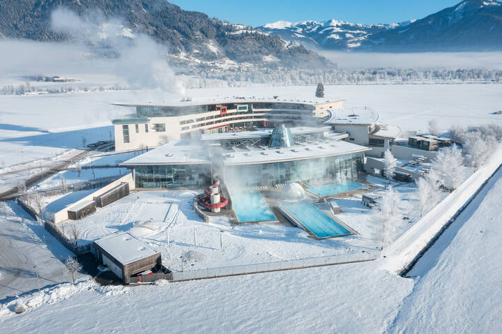 Aerial view of the wintery Tauerna Spa thermal baths with water slides and steam clouds, surrounded by snow and mountains. | © TAUERN SPA Zell am See-Kaprun