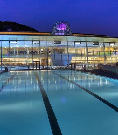 Modern spa building at night with illuminated pool in the foreground, glass facade and colorful lights, peaceful atmosphere. | © Photoart Reifmüller