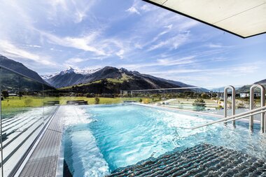 View of the outdoor pool at Tauern Spa with mountain scenery in the background, surrounded by mountains and clear blue sky. | © TAUERN SPA Zell am See-Kaprun