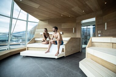 Two people relax in a sauna with large windows and wood interior in a spa. | © TAUERN SPA Zell am See-Kaprun