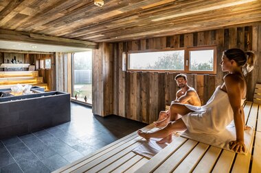 Two people relaxing in a sauna with wooden walls and large windows. | © TAUERN SPA Zell am See-Kaprun