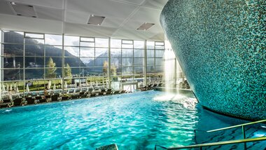 Indoor wellness and water park with a large pool, modern architecture, and large windows showing a scenic mountain landscape. | © TAUERN SPA Zell am See-Kaprun