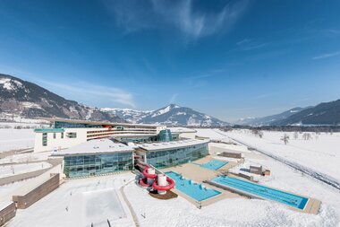 Aerial view of a winter hotel with indoor and outdoor swimming pools and water slide in a snowy landscape, surrounded by mountains. | © Alexander Maria Lohmann